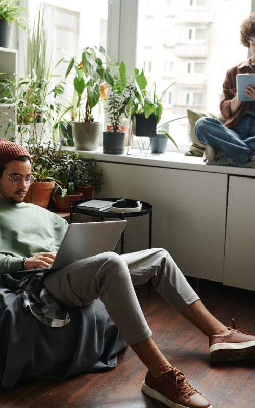 Young woman with tablet sitting on windowsill in front of male colleague with laptop networking in armchair during individual work