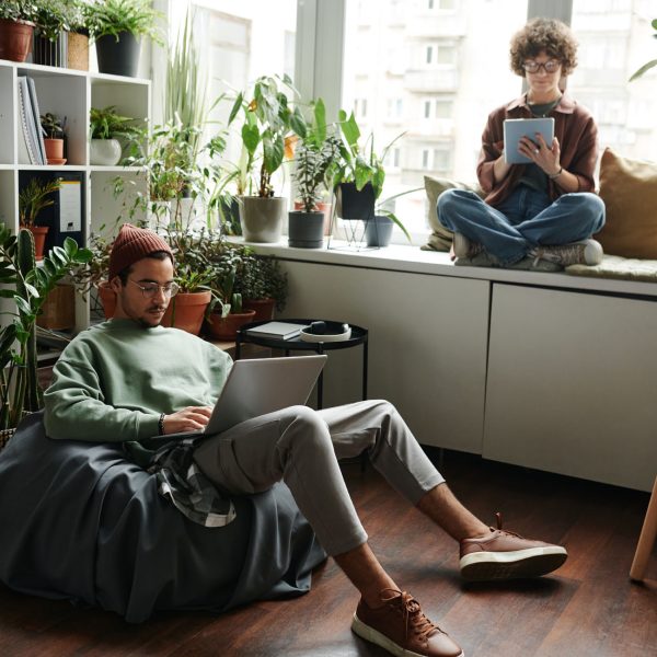 Young woman with tablet sitting on windowsill in front of male colleague with laptop networking in armchair during individual work